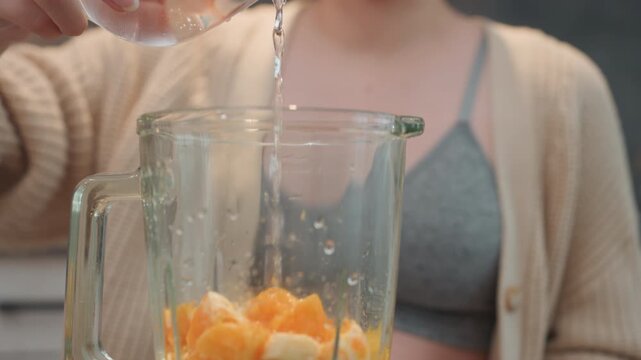 Pregnant woman adding water to fruit in blender jar, mandarin segments and banana visible, closeup pouring action with droplets on glass, focus on texture and hydration, expectant mother prepping