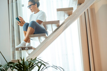 Woman in casual clothes sitting on modern stairs scrolling smartphone and smiling in bright home © simona