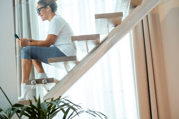 Fototapeta premium Woman in leggings sitting on modern staircase at home smiling while using smartphone in bright interior