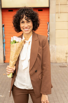 Woman in Brown Suit Holds Bouquet of Flowers on City Street