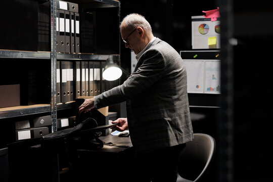 Old private investigator packing laptop up in suitcase after finishing investigation case. Senior detective in archive room office filled with confidential folders on bookshelves