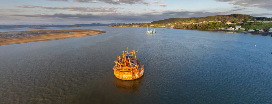 A large, rusty dredging barge sits in the calm waters of the Kawhia Harbour, Kawhia, Waikato, New Zealand, at sunrise. The barge is used for seabed maintenance and resource extraction.