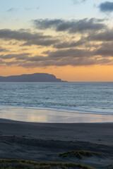 Obraz premium A serene sunset over the ocean at Kawhia Hot Water Beach, Kawhia, Waikato, New Zealand. The sky displays soft pastel colors as clouds drift by, reflecting on the wet sand.