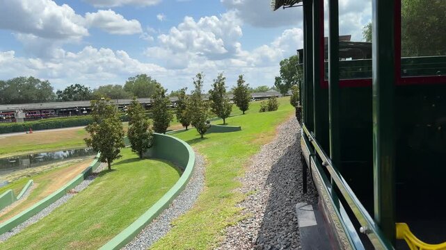 A vintage train travels through a green landscaped area under a blue sky. The scene shows scenic transportation, tourism, and relaxed travel.