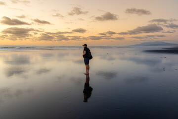 A lone man stands on a wet, sandy beach at dusk, gazing out at the ocean. The sky is filled with soft clouds, and the water reflects the fading light at Kawhia Hot Water Beach, Waikato, New Zealand. © Zenstratus