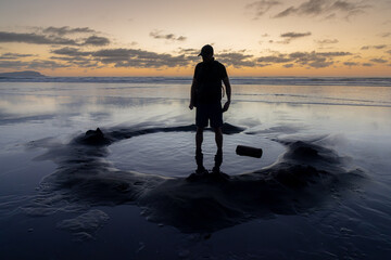 A man stands in thermal hot pool on a black sand beach at Kawhia Hot Water Beach, Kawhia, Waikato, New Zealand. © Zenstratus