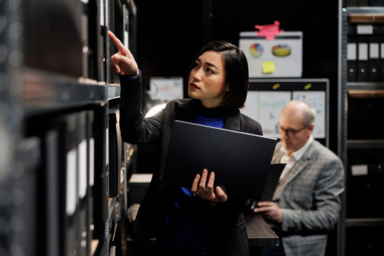 Asian private detective in criminal cases archive office looking for criminology file paperwork on cabinet shelves. Investigator woman surrounded by criminal case folders in agency repository room