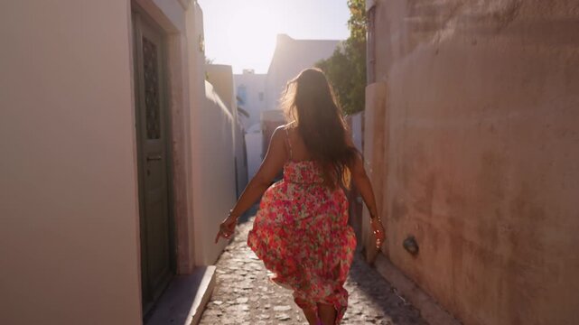 Back view of Young woman wearing colorful floral dress running through narrow sunlit alley between whitewashed buildings. Golden hour backlight creates warm glow on cobblestone path. Santorini, Greece