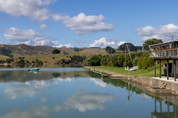 Obraz premium A tranquil scene unfolds at a waterfront building in Kawhia Harbour, Te Waitere, Waikato, New Zealand, with a small boat gliding across the calm water under a partly cloudy sky.