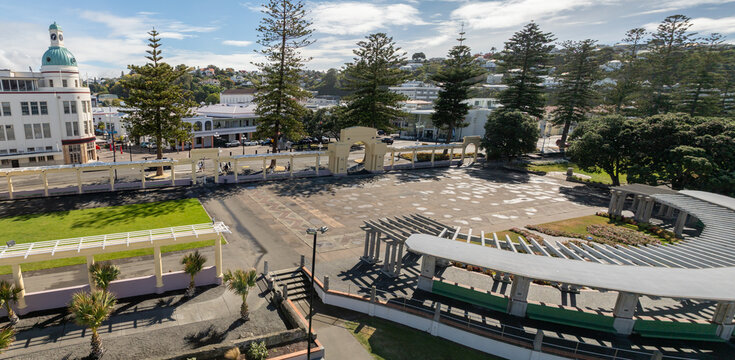 A sunny day in Napier, New Zealand, with people walking and cycling along the waterfront promenade. The iconic Art Deco buildings and lush greenery create a vibrant atmosphere. Marine Parade.