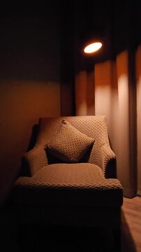 A cozy patterned armchair with a matching cushion sits under the warm glow of a modern pendant lamp, creating a dramatic and serene atmosphere with soft shadows against a dark curtain background.