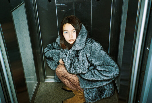 Person Squatting in an Elevator Wearing a Large Coat and Boots
