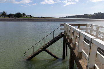 Fototapeta premium A wooden pier with stairs leading into the water at a beach in Raglan, Waikato, New Zealand. The scene captures the serene coastal landscape.