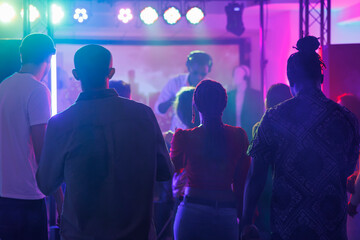 Fototapeta premium People partying and attending dj performance at nightclub discotheque. Young diverse men and women standing on crowded dark dancefloor while musician playing on stage in club