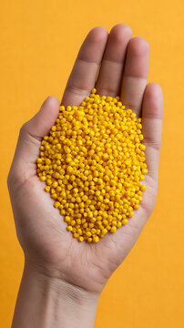 Close up view of a human hand holding a handful of small yellow plastic pellets or granules used for industrial manufacturing and production, set against a matching yellow background.