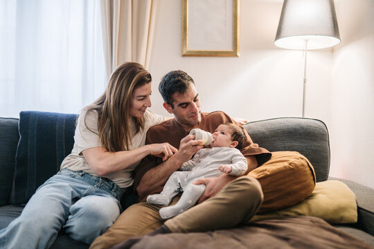 Parents feeding baby bottle at home on sofa