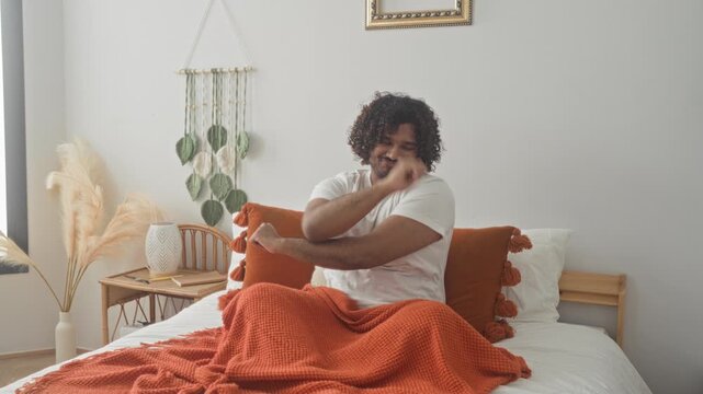 Young indian man on bed stretches arms and smiles with orange blanket and tassel pillow in studio; morning relaxation.