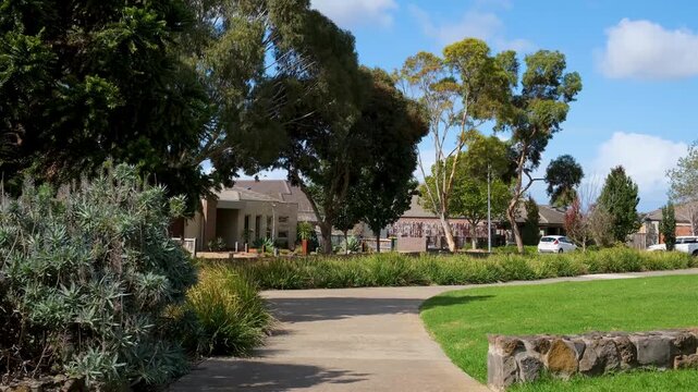 A winding concrete pathway, ornamental native plantings, manicured lawn, and residential homes. Tarneit suburban neighbourhood park with garden, footpath and houses, Melbourne Australia