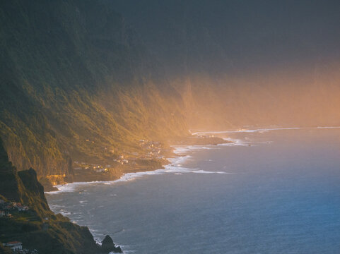 Aerial view of the rugged coastline with steep green cliffs and small coastal villages during golden hour in Madeira, Portugal.