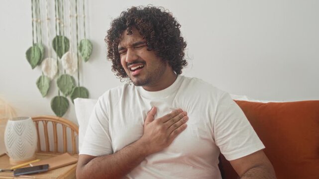 Young indian man clutching chest and pressing hand to chest on bed in studio with bedside table and smartphone, suggesting heartattack; pain.