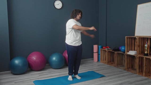 Man stretching on yoga mat in studio with exercise balls and wall clock, white t shirt and navy pants, arm raised in side stretch; serenity focus.
