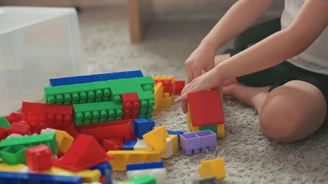 Boy building colorful blocks on carpet, focused solo play session with bright plastic pieces scattered across floor, small hands stacking red and yellow bricks, cozy indoor light, sensory learning