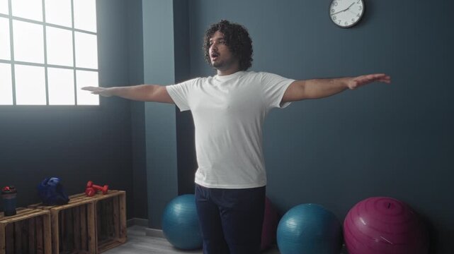 Man in white t-shirt, arms outstretched, balancing near blue and pink exercise balls and red dumbbells on wooden crates beside a large window and clock in a studio; calm focus balance.