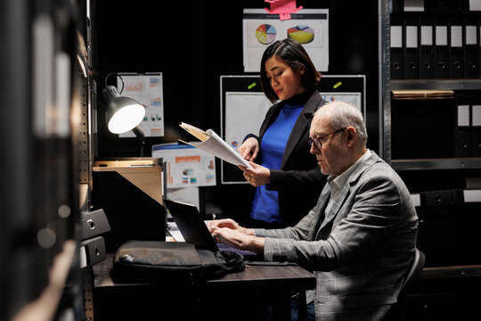 Professional administration employees imputing bookkeeping administrative data paperwork on laptop. Accountancy archival cabinet room filled with folders and diagram charts