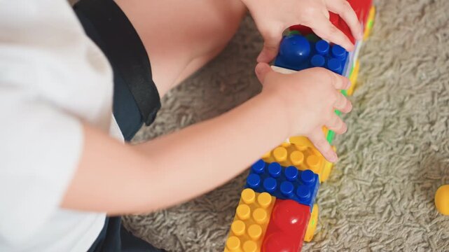 Toddler son playing with colorful blocks, focused stacking large plastic bricks on soft carpet, hands guiding blue red yellow pieces, closeup of fine motor practice and early learning exploration