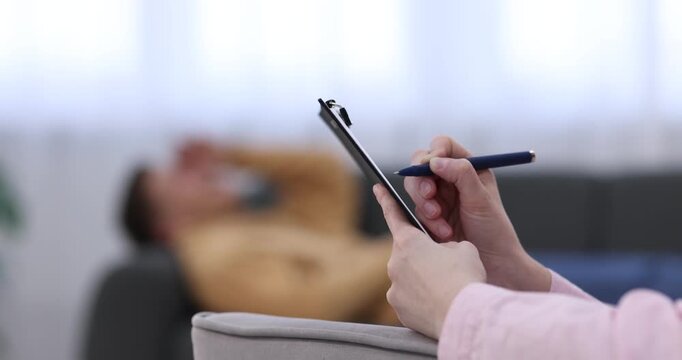 Professional psychologist making notes on clipboard during therapy session with patient indoors, selective focus