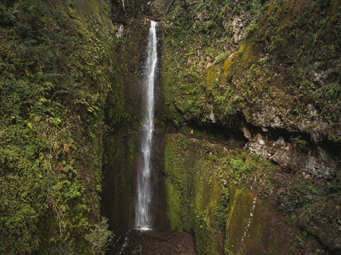 Aerial view of a tall waterfall cascading down a lush green cliff with a narrow walking path in Madeira, Portugal.