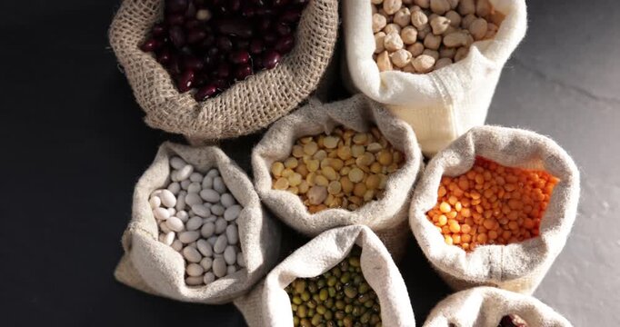 Different types of legumes in burlap bags on black table, flat lay. Camera moving right