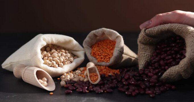 Woman pouring dried red beans from burlap bag and other legumes at black table, closeup
