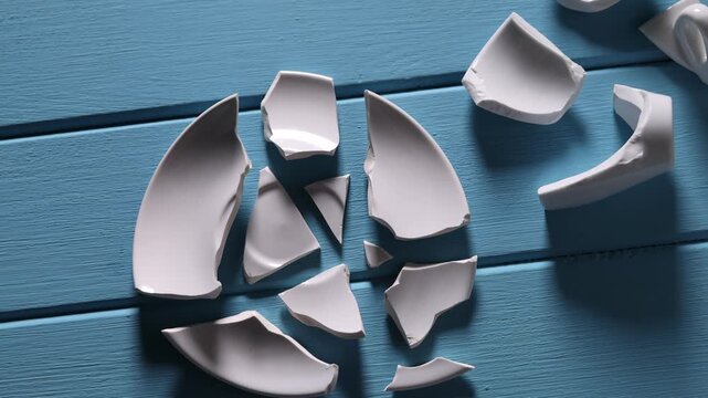Pieces of broken saucer and cup on light blue wooden table, flat lay. Camera moving