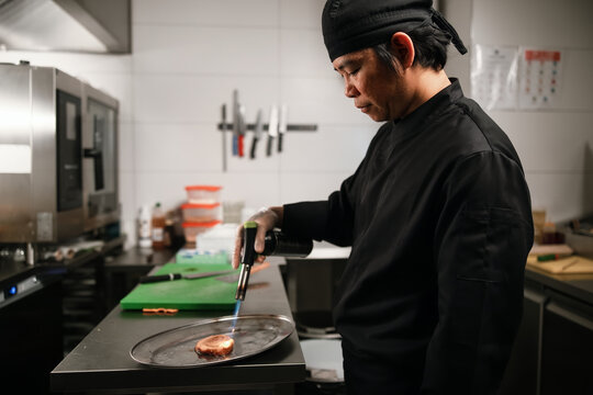 Professional Chef Searing Meat With Culinary Torch In Kitchen