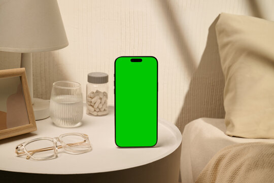 Green screen smartphone on bedside table with glasses and medication