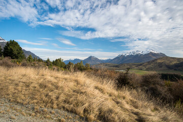 Fototapeta premium Paysage de prairie dorée aride avec montagnes enneigées et ciel bleu nuageux, région de Wanaka, île du Sud, Nouvelle-Zélande, automne