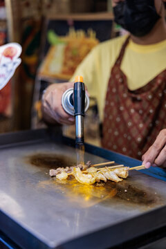 Street vendor torches skewers of meat and seafood.