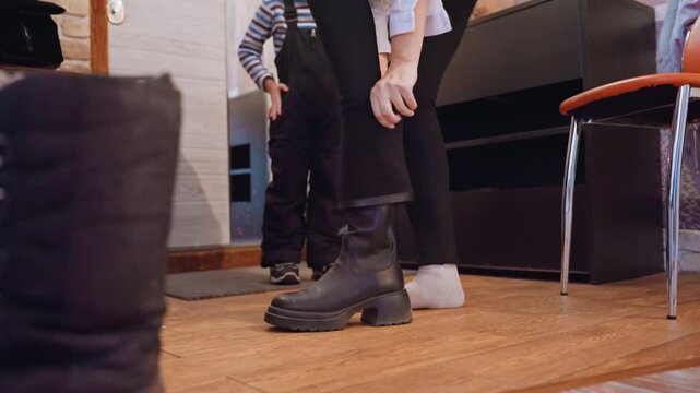 Mom and son putting on boots in cozy entryway, mother kneeling to fasten leather boot while young boy in socks waits on hardwood floor beside chair, warm morning light, intimate candid moment of care