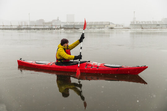 Profile of Kayaker Paddling Red Boat in Icy Urban Harbor