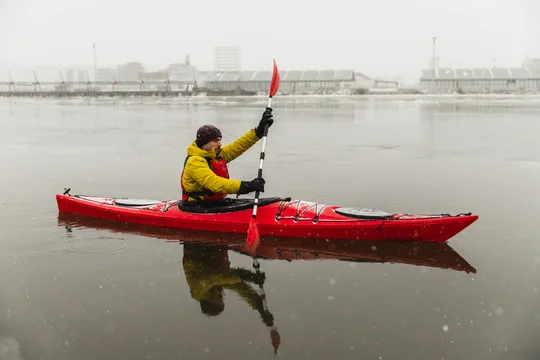 Profile of Kayaker Paddling Red Boat in Icy Urban Harbor