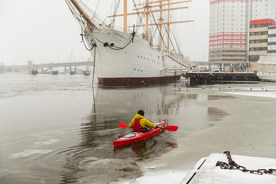 Red Kayak Paddling Towards Large Sailing Ship in Icy Harbor