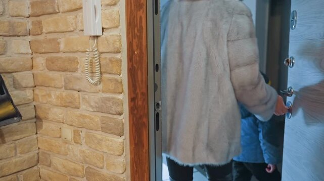 Wide shot mom in fur coat opening door into hallway, hand on silver knob, brick wall and intercom visible, brief glimpse of child in blue jacket, morning departure mood, practical routine, soft indoor