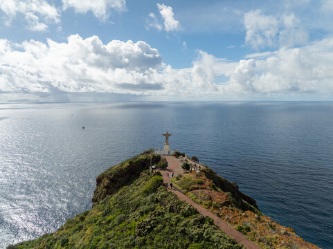 Aerial view of the Cristo Rei statue standing on a rocky cliff overlooking the vast blue Atlantic Ocean with a walking path and visitors in Canico, Madeira, Portugal.