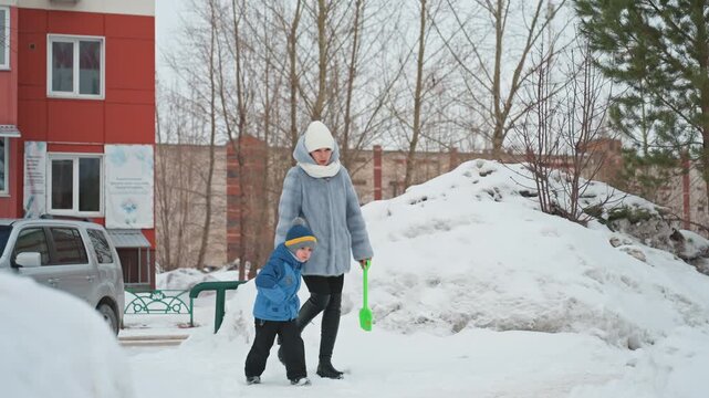 Mom and son walking in snow, mother guiding child across snowy courtyard by red apartment building, bundled in blue coat and knit hat, child investigates snow pile with playful curiosity while mom