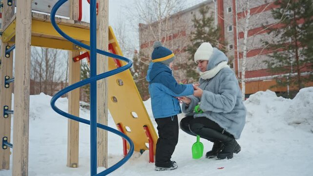 White mom kneels to help son adjust mittens at snowy playground, mom in warm gray coat and white beanie carefully fixes child zipper, green shovel rests on snow near spiral climber, intimate winter