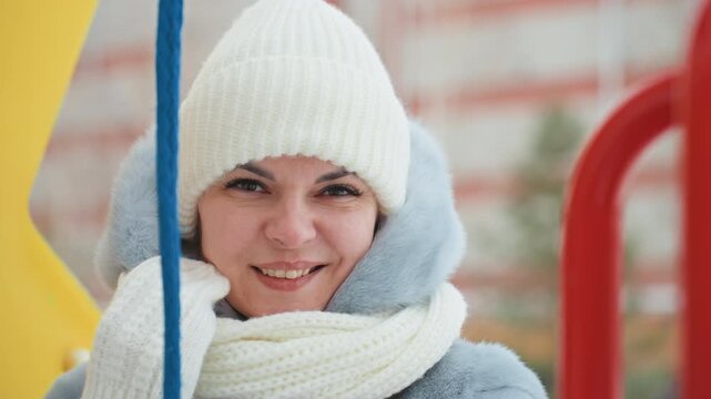 White mom bundled in winter coat at playground, smiling behind colorful slide rails, wearing knit beanie and thick scarf, rosy cheeks from cold, candid portrait conveying cozy maternal warmth