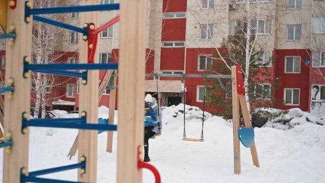 Snowy courtyard mom and son walking, apartment building backdrop, colorful swing set and climbing frame, mother supervising child bundled in coat and hat, footprints in fresh snow, overcast light,