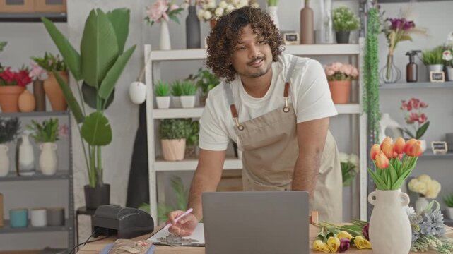Man florist in apron leaning and writing on a clipboard beside laptop at a flower shop counter in a building; warmth friendly service.