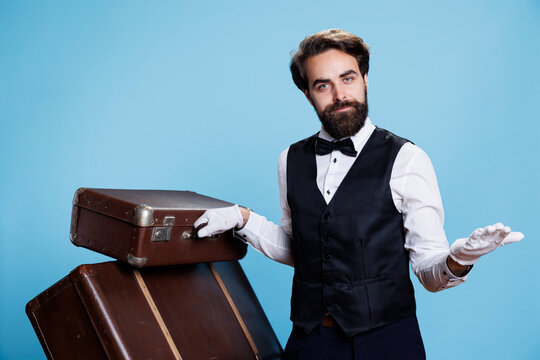 Luxurious hotel porter with luggage posing over blue background to advertise professional hospitality industry. Man working as bellhop with formal suit and white gloves, gracious doorman.